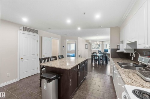 Kitchen featuring a large island with stone countertop, dark wood-finish cabinetry, white upper and lower cabinetry, and tile flooring - #104 10221 111 St Nw, Edmonton, AB - Indoor Photo Showing Kitchen With Double Sink