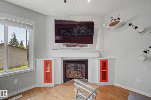 Living area featuring a corner fireplace with a white mantel, built-in shelving with red accents, a large window, and wood-finish flooring - 9935 155 Street Nw, Edmonton, AB - Indoor With Fireplace