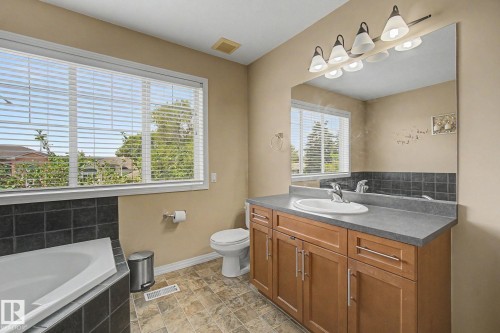 Corner bathtub with black subway tile surround, wood-finish vanity with an integrated sink and gray countertop, large window with blinds, and patterned tile flooring - 9935 155 Street Nw, Edmonton, AB - Indoor Photo Showing Bathroom