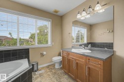 Corner bathtub with black subway tile surround, wood-finish vanity with an integrated sink and gray countertop, large window with blinds, and patterned tile flooring - 
