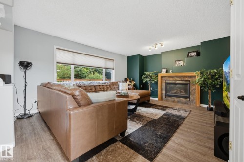 Living area featuring wood-finish flooring, a large window, a fireplace with a stone surround, and track lighting - 15 Orchid Place, St. Albert, AB - Indoor Photo Showing Living Room With Fireplace