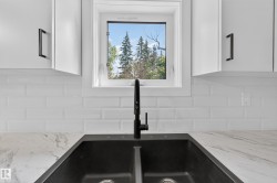 Living Space # 2  Dual-basin sink with a matte black faucet, complemented by white subway tile backsplash, white cabinetry with black hardware, a window overlooking trees, and light-toned, veined countertops - 