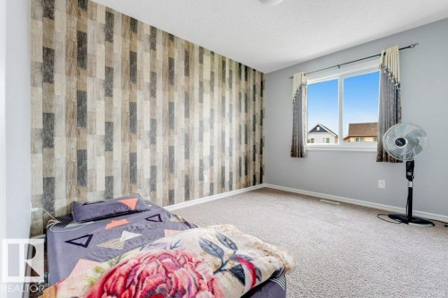 Spacious bedroom with neutral carpeting, large windows with natural light, vertical striped accent wall featuring earth-tone finishes, and standalone pedestal fan - 2436 18 Avenue Nw, Edmonton, AB 