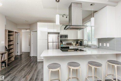 Modern kitchen featuring white cabinetry, stainless steel appliances, a white countertop island with seating, a tile backsplash, and wood-finish flooring - #208 9907 91 Av Nw, Edmonton, AB - Indoor Photo Showing Kitchen With Upgraded Kitchen