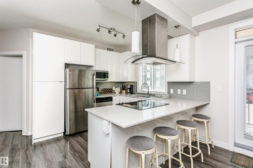 Modern kitchen featuring an extended island with a built-in cooktop, stainless steel appliances, white flat-panel cabinetry, a stainless steel range hood, and wood-finish flooring - #208 9907 91 Av Nw, Edmonton, AB - Indoor Photo Showing Kitchen With Stainless Steel Kitchen With Upgraded Kitchen