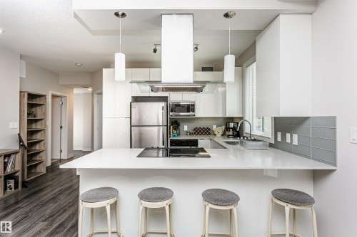 Modern kitchen featuring a white countertop with integrated cooktop, stainless steel appliances, white cabinetry, and a grey tile backsplash - #208 9907 91 Av Nw, Edmonton, AB - Indoor Photo Showing Kitchen With Stainless Steel Kitchen With Upgraded Kitchen