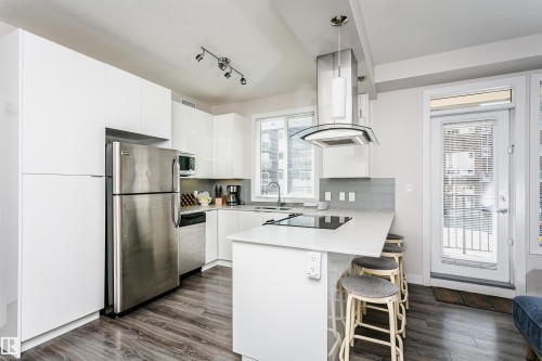 Modern kitchen featuring white cabinetry, stainless steel appliances, a white island with seating, a glass-panel door, and wood-finish flooring - #208 9907 91 Av Nw, Edmonton, AB - Indoor Photo Showing Kitchen With Stainless Steel Kitchen With Upgraded Kitchen