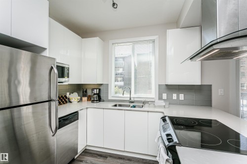 Contemporary kitchen featuring white cabinetry, stainless steel appliances, a grey tile backsplash, a curved countertop, and wood-finish flooring - #208 9907 91 Av Nw, Edmonton, AB - Indoor Photo Showing Kitchen With Double Sink