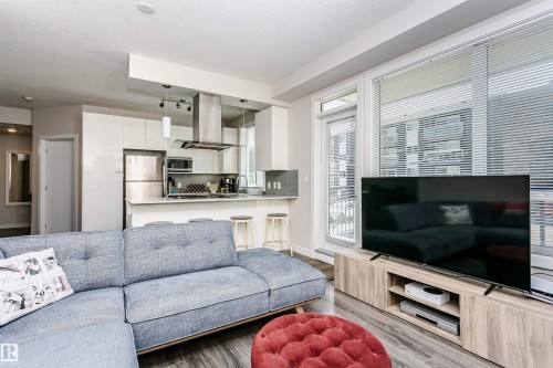 Open-concept living space featuring wood-finish flooring, a contemporary kitchen with stainless steel appliances, white flat-panel cabinetry, a breakfast bar, and large windows with blinds - #208 9907 91 Av Nw, Edmonton, AB - Indoor Photo Showing Living Room