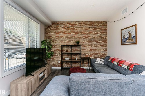 Living area featuring an exposed brick accent wall, wood-finish flooring, and large windows with horizontal blinds - #208 9907 91 Av Nw, Edmonton, AB - Indoor Photo Showing Other Room