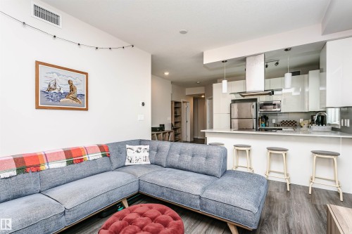 Open-concept living space with wood-finish flooring, a kitchen island with seating, stainless steel appliances, and sleek white cabinetry - #208 9907 91 Av Nw, Edmonton, AB - Indoor Photo Showing Living Room