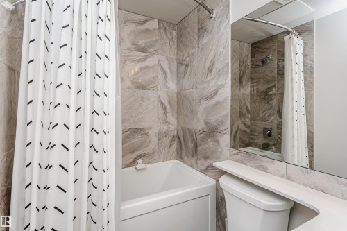 Bathroom featuring a white bathtub, full-height tile surround, wall-mounted mirror, and a white toilet with an integrated shelf - #208 9907 91 Av Nw, Edmonton, AB - Indoor Photo Showing Bathroom