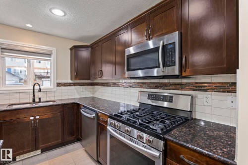 Kitchen featuring dark wood-finish cabinetry, stainless steel appliances, a gas range, dark speckled countertops, and a white subway tile backsplash with a decorative accent band - 1209 30 Avenue Nw, Edmonton, AB - Indoor Photo Showing Kitchen With Double Sink With Upgraded Kitchen