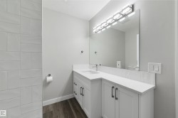 White subway tile shower surround, L-shaped vanity with integrated sink, white countertop, dark wood-finish flooring, and a large wall-mounted mirror with an overhead light fixture - 