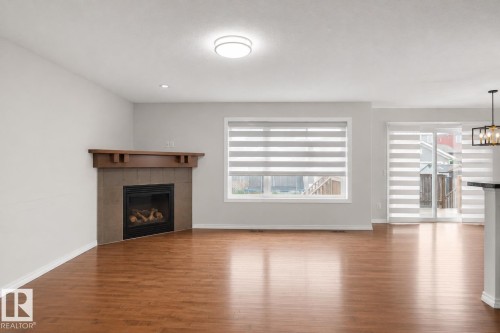 Wood-finish flooring throughout, featuring a tiled fireplace with wood mantel, large window with blinds, and sliding glass doors with blinds - 8432 18 Avenue Sw, Edmonton, AB - Indoor Photo Showing Living Room With Fireplace