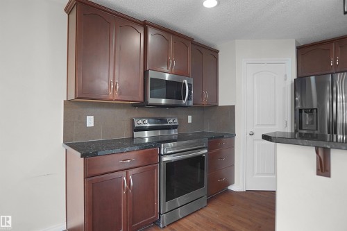 Kitchen featuring dark wood cabinetry, stainless steel appliances, dark stone countertops, and a tile backsplash - 8432 18 Avenue Sw, Edmonton, AB - Indoor Photo Showing Kitchen