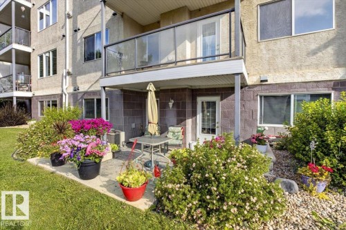 Ground-level patio with stone pavers, a balcony featuring glass railings, and a manicured lawn area - #116 237 Youville Dr E Nw, Edmonton, AB - Outdoor