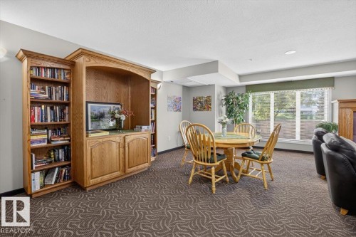 Built-in wooden cabinetry with open shelving and closed storage, patterned carpet flooring, large windows, and recessed ceiling lighting - #116 237 Youville Dr E Nw, Edmonton, AB - Indoor