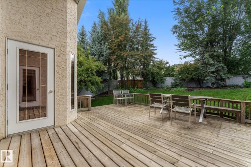 Expansive wood deck with an integrated bench, featuring a glass-paneled door, curved architectural wall, and surrounding green space with mature trees - 8 Osborne Close, St. Albert, AB - Outdoor