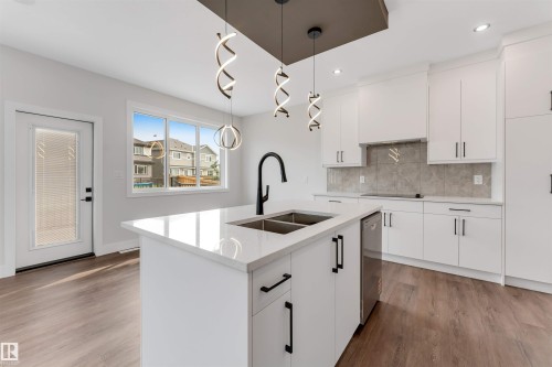 Modern kitchen featuring a central island with an undermount sink, white cabinetry, integrated dishwasher, and wood-finish flooring - 329 Balsam Link, Leduc, AB - Indoor Photo Showing Kitchen With Double Sink With Upgraded Kitchen