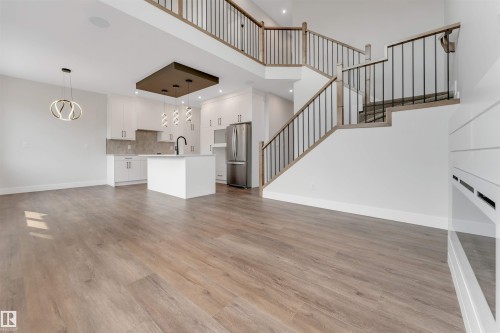 Open-concept living space featuring wood-finish flooring, a contemporary kitchen with white cabinetry, a large kitchen island, stainless steel appliances, and a modern staircase with dark metal spindles and wood handrails - 329 Balsam Link, Leduc, AB - Indoor Photo Showing Other Room