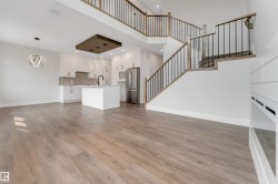 Open-concept living space featuring wood-finish flooring, a contemporary kitchen with white cabinetry, a large kitchen island, stainless steel appliances, and a modern staircase with dark metal spindles and wood handrails - 