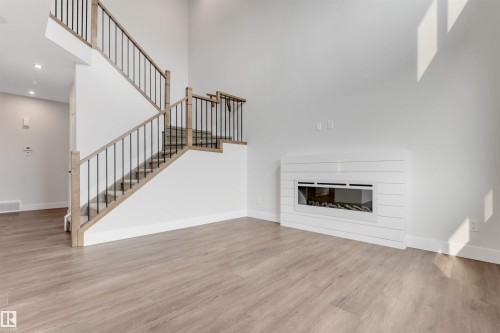 Contemporary living area featuring a modern linear fireplace with a white shiplap-style surround, two-story ceilings, wood-finish flooring, and a staircase with light wood newel posts and black metal spindles - 329 Balsam Link, Leduc, AB - Indoor With Fireplace