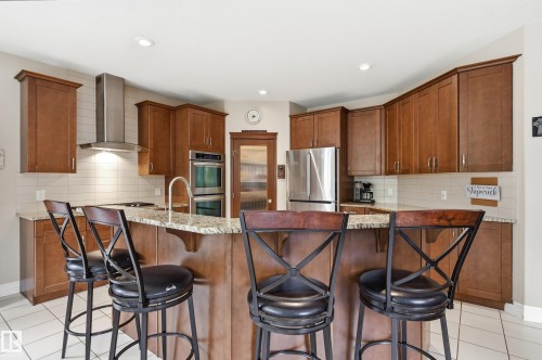 Kitchen featuring wood cabinetry, stainless steel appliances, a tiled backsplash, and an island with a stone countertop - 2416 Warry Place Sw, Edmonton, AB - Indoor Photo Showing Kitchen