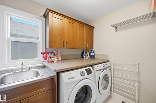 Dedicated laundry room featuring side-by-side front-load washers and dryers, wood cabinetry with ample storage, stainless steel sink with chrome faucet, quartz countertop workspace, and natural light from a double-hung window - 2416 Warry Place Sw, Edmonton, AB - Indoor Photo Showing Laundry Room