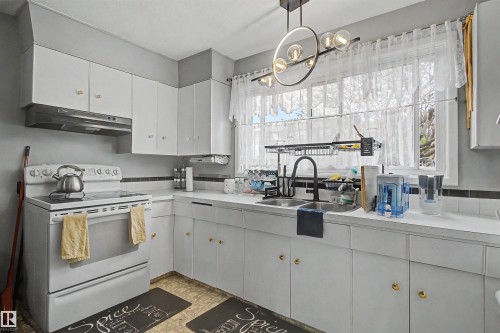 White cabinetry with gold-finish hardware, recessed kitchen sink with gooseneck faucet, light wood-finish flooring, and a contemporary chandelier - 5724 143 Avenue Nw, Edmonton, AB - Indoor Photo Showing Kitchen With Double Sink