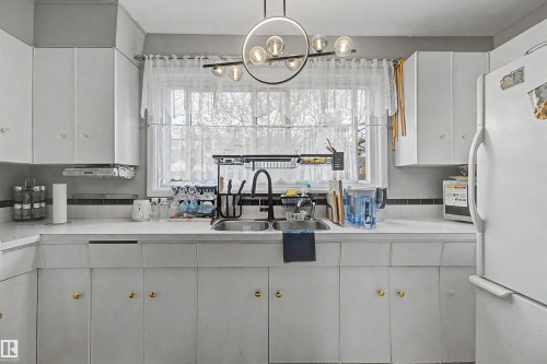 White cabinetry with gold-tone hardware, a double basin sink with a matte black faucet, and a contemporary linear chandelier with globe lights - 5724 143 Avenue Nw, Edmonton, AB - Indoor Photo Showing Kitchen With Double Sink
