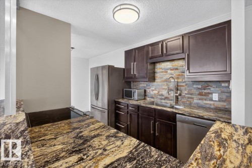 Kitchen featuring dark wood cabinetry, a stacked stone backsplash, stainless steel appliances, and patterned countertops - #506 12121 Jasper Av Nw, Edmonton, AB - Indoor Photo Showing Kitchen With Double Sink
