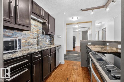 Kitchen featuring dark wood cabinetry, a stacked stone backsplash, stone-finish countertops, stainless steel appliances, and wood-finish flooring - #506 12121 Jasper Av Nw, Edmonton, AB - Indoor Photo Showing Kitchen With Upgraded Kitchen