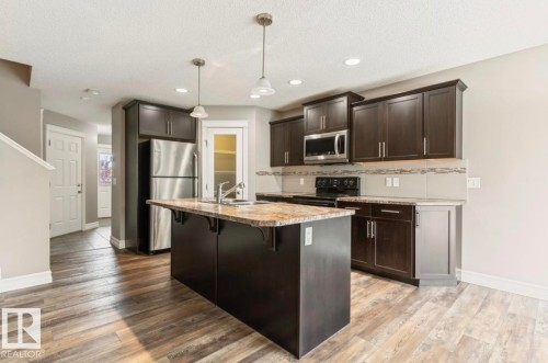 Kitchen featuring dark wood-finish cabinetry, stainless steel appliances, a center island with a sink, pendant lighting, and wood-finish flooring - 79 Catalina Crest, Fort Saskatchewan, AB - Indoor Photo Showing Kitchen With Upgraded Kitchen