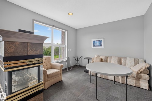Interior space featuring a large window, a built-in fireplace with a decorative mantel, neutral gray wall paint, recessed lighting, and gray patterned carpet flooring - #105 10421 42 Av Nw, Edmonton, AB - Indoor Photo Showing Living Room With Fireplace