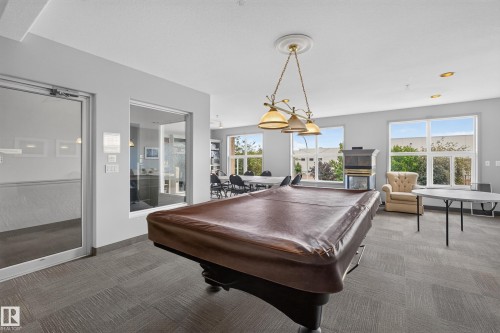 Spacious common area featuring a covered billiards table, fireplace, numerous windows, and grey-toned carpet tile flooring - #105 10421 42 Av Nw, Edmonton, AB - Indoor Photo Showing Other Room