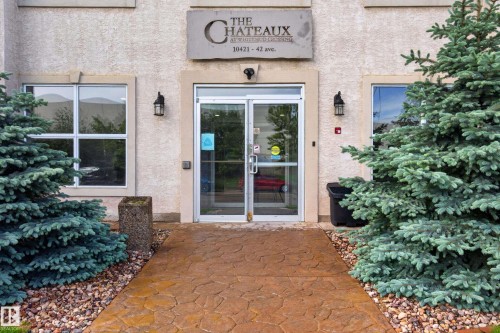 Front entrance featuring a stamped concrete walkway, mature evergreen plantings, and a sign with prominent building identification - #105 10421 42 Av Nw, Edmonton, AB - Outdoor