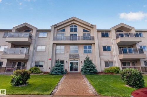 Multi-story residential building featuring balconies with glass railings, an arched central entrance, and a paved walkway - #105 10421 42 Av Nw, Edmonton, AB - Outdoor With Balcony With Facade