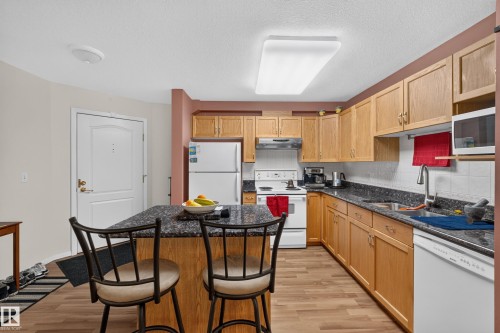 Kitchen featuring wood-finish flooring, an island with dark granite-style countertop, light wood cabinetry, a white tile backsplash, and a white appliance suite - #105 10421 42 Av Nw, Edmonton, AB - Indoor Photo Showing Kitchen