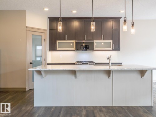 Kitchen island with a light-colored countertop, dark wood-finish cabinetry, built-in gas range, stainless steel sink with a gooseneck faucet, and three pendant lights - 97 Astoria Point(E), Devon, AB - Indoor Photo Showing Kitchen With Upgraded Kitchen