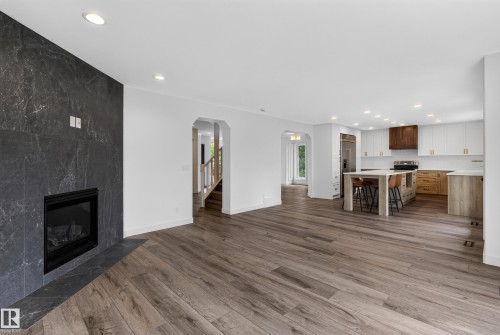 Expansive wood-finish flooring, a modern fireplace with a dark stone surround, recessed lighting, and a kitchen featuring a large island with seating, built-in stainless steel appliances, and two-tone cabinetry - 229 Hillcrest Crest, Sherwood Park, AB - Indoor Photo Showing Living Room With Fireplace