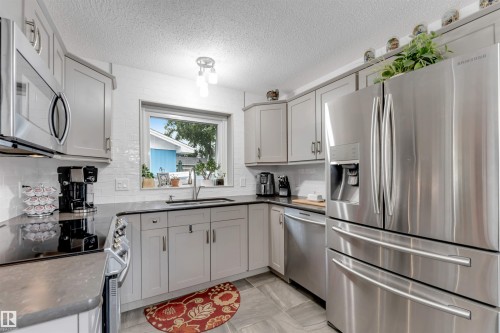 Modern kitchen featuring light gray cabinetry, stainless steel appliances, a white subway tile backsplash, dark countertops, and tile flooring - 6008 37 Avenue Nw, Edmonton, AB - Indoor Photo Showing Kitchen