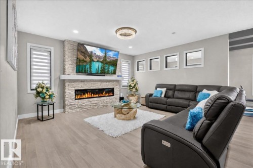 Light wood-finish flooring, stone-clad fireplace with mantel, recessed electric fireplace, multiple windows with blinds, and a decorative ceiling fixture - 6813 Knox Loop Sw, Edmonton, AB - Indoor Photo Showing Living Room With Fireplace