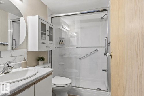 Walk-in shower with sliding glass door, white vanity with integrated sink, chrome faucet, wall-mounted storage cabinet, and round mirror - #102 6710 158 Av Nw, Edmonton, AB 