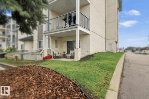 Covered patio with an exterior sliding door, an upper-level balcony with metal railing, and an adjacent landscaped area with bark mulch and grass - #102 6710 158 Av Nw, Edmonton, AB 
