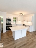 Modern kitchen featuring a central island with a white countertop, white shaker-style cabinetry, stainless steel appliances, wood-finish flooring, and a contemporary ceiling fixture - 