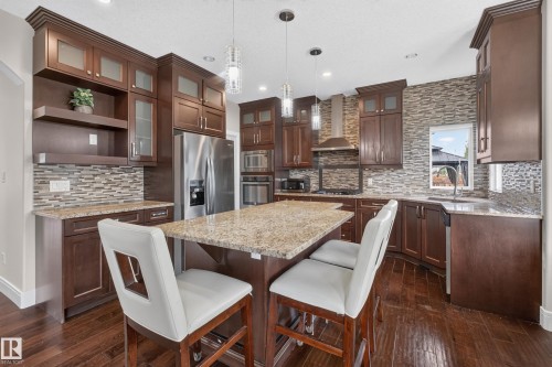 Kitchen featuring rich wood cabinetry, granite countertops, a central island, stainless steel appliances, and wood-finish flooring - 7014 Chivers Loop Sw, Edmonton, AB 