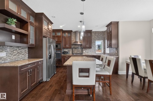 Gourmet kitchen featuring dark wood cabinetry, stainless steel appliances, granite countertops, a mosaic tile backsplash, and wood-finish flooring - 7014 Chivers Loop Sw, Edmonton, AB 