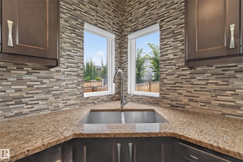 Corner sink with a gooseneck faucet, dual stainless steel basins, granite countertops, and mosaic tile backsplash, complemented by dark wood-finish cabinetry - 7014 Chivers Loop Sw, Edmonton, AB 