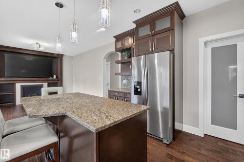 Built-in entertainment center with fireplace, stainless steel refrigerator, dark wood-finish cabinetry, granite countertops, and wood-finish flooring - 7014 Chivers Loop Sw, Edmonton, AB 